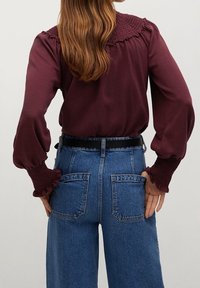 Bordeaux blouse met geplooide schouders en gerimpelde manchetten, gecombineerd met hoge taille blauwe denim jeans met achterzakken.