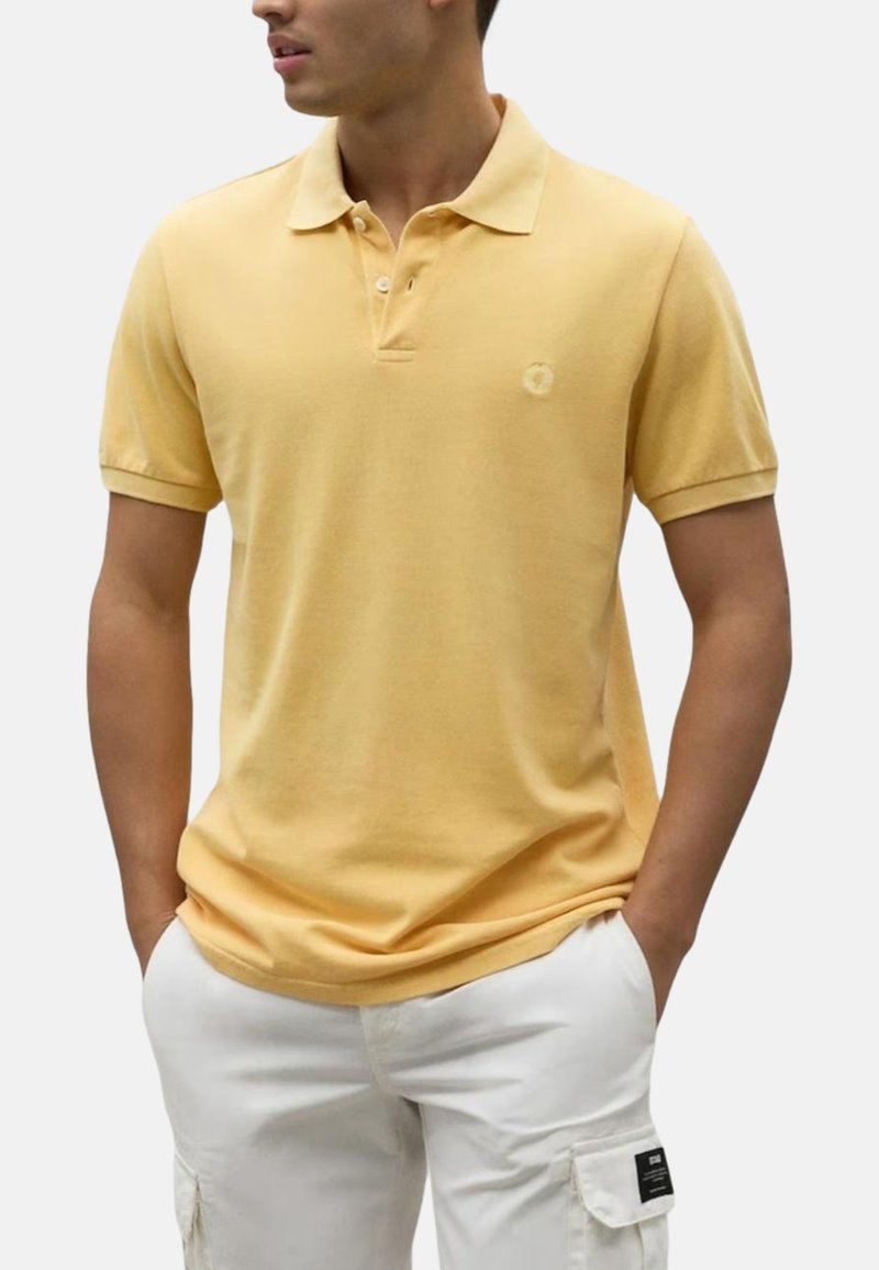 Camisa polo de algodón amarilla con cuello, tapeta de tres botones y pequeño logo en el pecho, combinada con pantalones cortos cargo blancos.