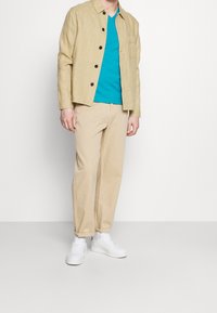 Béžová lněná bunda, tyrkysové tričko s výstřihem do V a světle béžové kalhoty. Outfit doplňují bílé tenisky. Jednoduchý design, uvolněný střih.