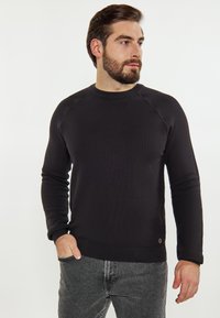 Zwarte gebreide trui met een ronde halslijn en lange mouwen. Heeft ribgebreide manchetten en zoom, textuurstof en subtiele branding aan de onderkant.