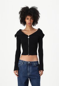 Čierny rebrovaný crop top s off-shoulder dizajnom, dlhými rukávmi a predným zipsom. Uložený s vyblednutými modrými džínsami s vysokým pásom.