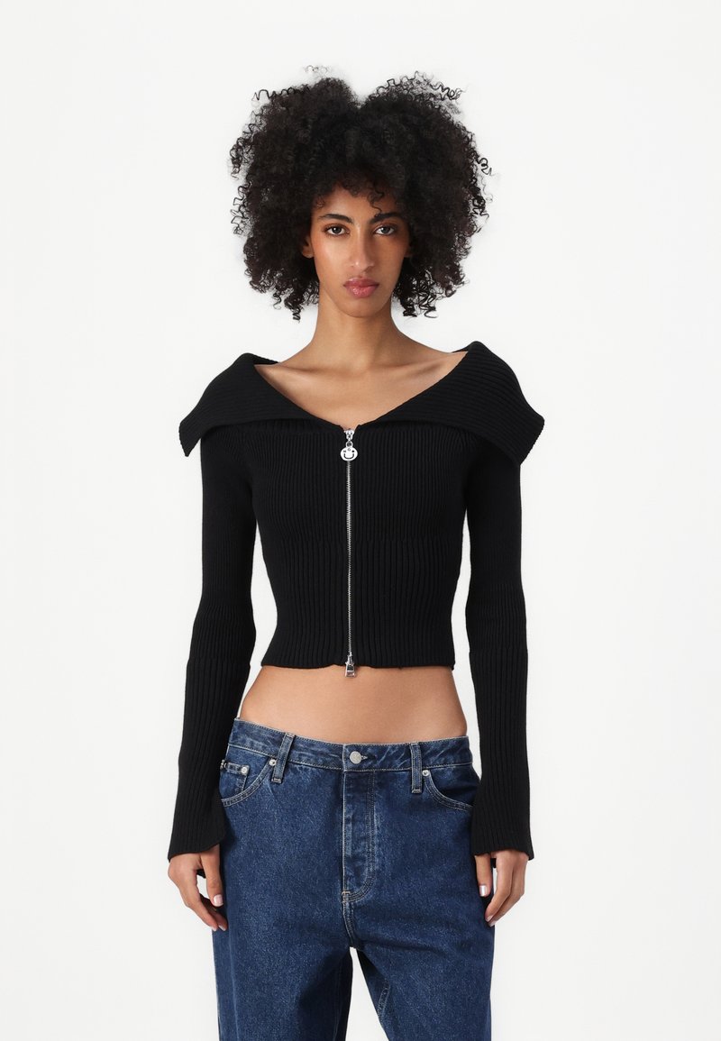 Čierny rebrovaný crop top s off-shoulder dizajnom, dlhými rukávmi a predným zipsom. Uložený s vyblednutými modrými džínsami s vysokým pásom.