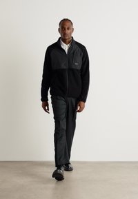 Svart zip-up fleecejakke med en grå frontpanel, kombinert med mørke bukser og svarte sko. Glatt tekstur, ingen synlige mønstre eller logoer.