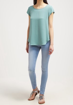 Femme portant un haut ample vert clair à manches courtes, un jean skinny bleu clair et des sandales argentées ornées, debout devant un fond uni.