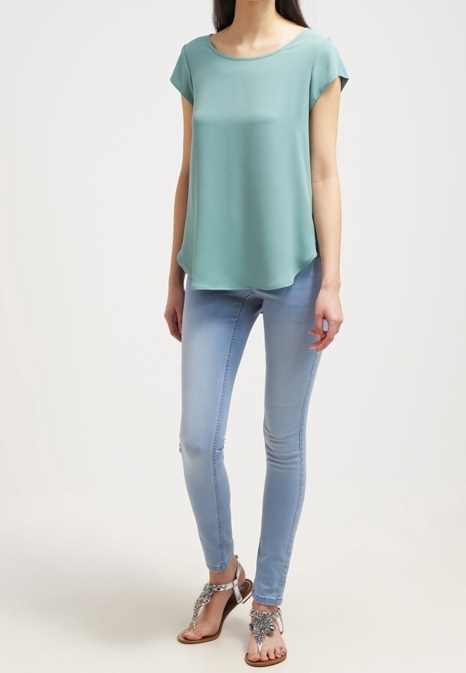 Femme portant un haut ample vert clair à manches courtes, un jean skinny bleu clair et des sandales argentées ornées, debout devant un fond uni.
