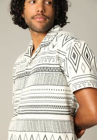 Korte mouwen shirt in het wit en zwart, met geometrische patronen van lijnen en diamanten. De stof lijkt textuur te hebben met een kraagdetail.