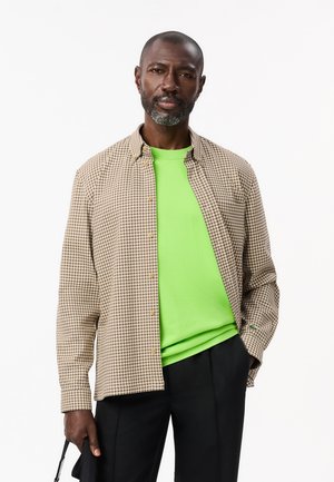 Camisa de cuadros en tonos marrones con cierre de botones sobre una camiseta de color verde neón. Pantalones negros completan el atuendo. Estructura de tejido simple y ligero.