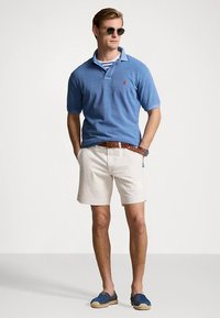 Niebieska koszulka polo z krótkim rękawem, jasnobeżowe szorty, brązowy pleciony pasek oraz niebieskie espadryle; nosi okulary przeciwsłoneczne.