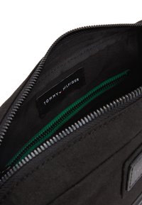 Borsa in tessuto nero con chiusura a zip, dotata di una tasca interna verde e un'etichetta Tommy Hilfiger. Texture liscia e dettagli di cucitura sottili.