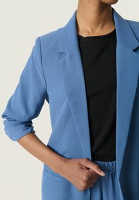 Modrý blejzr s klopovými límci, předními kapsami a ohrnutými rukávy; nošený přes černý top s kulatým výstřihem a elastickým pasem.