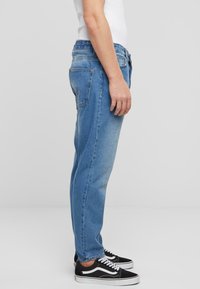 Jeans de denim azul claro con un corte relajado, que incluyen bolsillos laterales, bolsillos traseros de parche y un leve desgaste. Llevados con zapatillas negras.