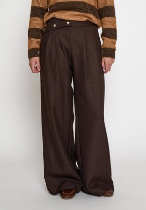 Persona che indossa pantaloni marrone scuro a vita alta e gamba larga con cintura abbottonata e maglione marrone a righe, in piedi su sfondo bianco.
