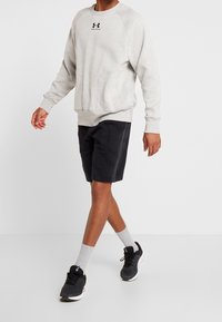 Sudadera gris con textura moteada, logo negro, combinada con pantalones cortos negros, calcetines grises y zapatillas deportivas negras. Postura caminando.