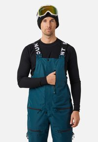 Uomo che indossa pantaloni da neve color verde acqua, maglia nera a maniche lunghe, berretto nero e occhiali da sci, con la mano nella tasca sul petto.