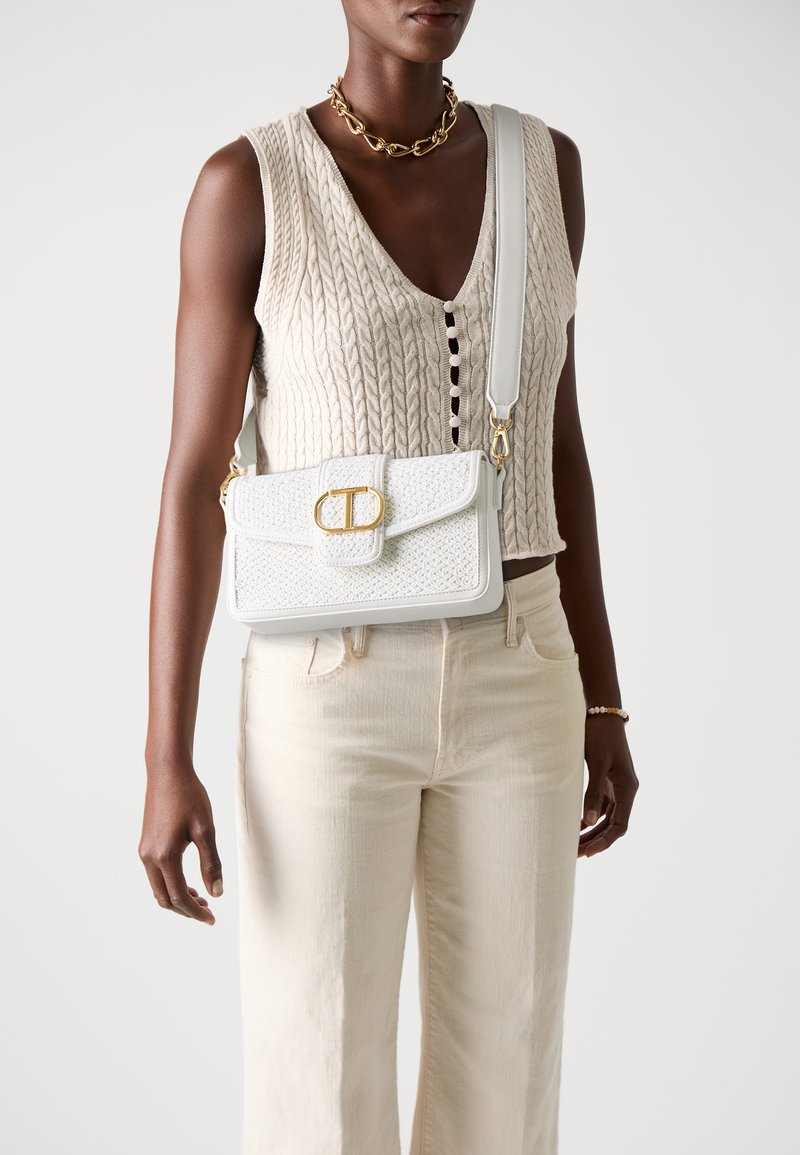 Sac blanc texturé avec quincaillerie dorée, de forme rectangulaire, avec une fermeture à rabat. Porté en travers du corps avec un haut en tricot léger et un jean clair.
