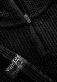 Cappotto in pile nero con zip e texture a coste. Presenta un piccolo etichetta con la scritta "ISCRIVITI" e un testo aggiuntivo dettagliato.
