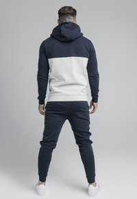 Felpa color blu navy e grigia a blocchi di colore con maniche lunghe e cappuccio, abbinata a pantaloni jogger coordinati e sneakers bianche. Tessuto strutturato.