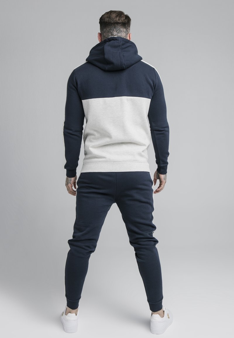 Felpa color blu navy e grigia a blocchi di colore con maniche lunghe e cappuccio, abbinata a pantaloni jogger coordinati e sneakers bianche. Tessuto strutturato.