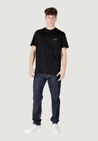 Zwarte katoenen T-shirt met korte mouwen en een ronde hals, voorzien van een klein gouden "A/X" logo op de borst. Gepaard met donkere jeans en witte sneakers.