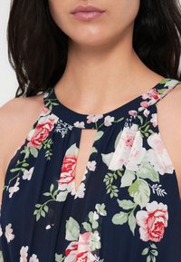 Femme portant une robe bleu marine sans manches avec un motif floral comprenant des fleurs roses et rouges et un décolleté en trou de serrure.