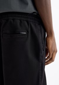 Pantalon de jogging noir avec taille élastique, poche zippée à l'arrière et étiquette de marque discrète. Tissu lisse avec une coupe élégante et ajustée.