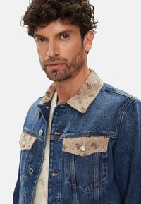 Chaqueta de mezclilla con cuello y bolsillos estampados en beige, con herrajes plateados y costuras en contraste, exhibiendo una combinación de colores azul y marrón claro.