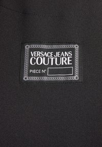 Black fabric label with white text reading "VERSACE JEANS COUTURE" and "PIECE N°," framed by a decorative rope-like border.