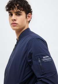 Νεαρός με σγουρά σκουρόχρωμα μαλλιά, φοράει μια ναυτικό μπλε μπουφάν μπόμερ με φερμουάρ σε τσέπη και λευκό κείμενο για οικολογική συνείδηση στο μανίκι.