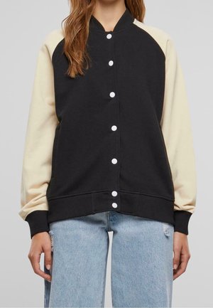 Jeune personne portant une veste varsity noire avec des manches crème et un jean bleu clair déchiré, debout devant un fond uni.