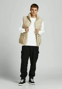 Jovem vestido com um colete acolchoado bege, sweat branco, calças cargo pretas e sapatilhas pretas com atacadores brancos, apoiado contra um fundo cinza.