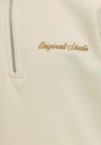 Krēmkrāsas zip-up džemperis ar gludu tekstūru, izšūtu "Originals Studio" zelta diegā un parasto rāvējslēdzēju.