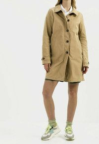 Hnědý voděodolný trenčkot s knoflíkovou légu, dvěma bočními kapsami a límcem, nošený přes šedé šortky a vícebarevné tenisky.