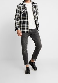 Svart och vit rutig skjorta med gula detaljer, kombinerad med en vit långärmad skjorta och mörkgrå jeans. Svarta läderskor med snörning.