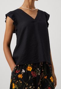 Blusa blu navy a pieghe con scollatura a V e maniche a coppetta arricciate, abbinata a una gonna nera a fantasia floreale. Tessuto morbido e vestibilità rilassata.