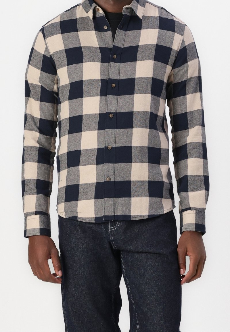 Man in een beige en marineblauwe grote geruite overhemd met donkerblauwe jeans, staand tegen een effen witte achtergrond.