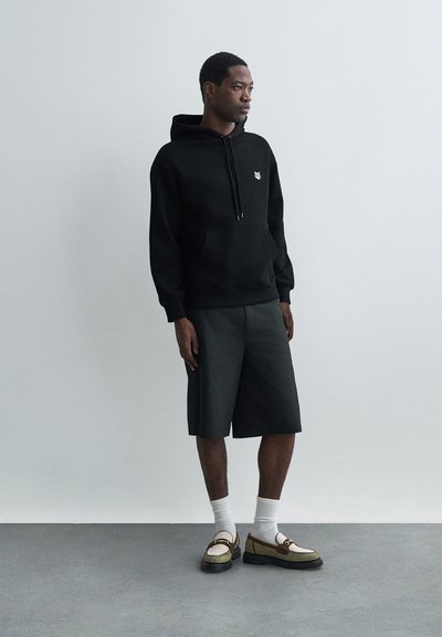 Sweat-shirt à capuche noir avec poche avant. Short gris, chaussettes blanches mi-mollet, et chaussures à enfiler avec des accents marron. Fond neutre.