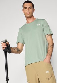 Camiseta de manga corta verde claro con cuello redondo y tela texturizada. Presenta un logo blanco en el lado izquierdo del pecho y se combina con pantalones cortos de carga color beige.