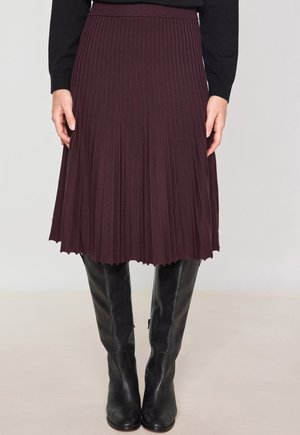 Jupe plissée en bourgogne profond, avec des plis verticaux et un ourlet festonné. Associée à des bottes noires ajustées au genou.