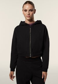 Haut cropped noir en mélange doux de coton, avec une fermeture éclair complète, un capuchon avec cordon de serrage et des poches latérales. Design simple sans motifs.