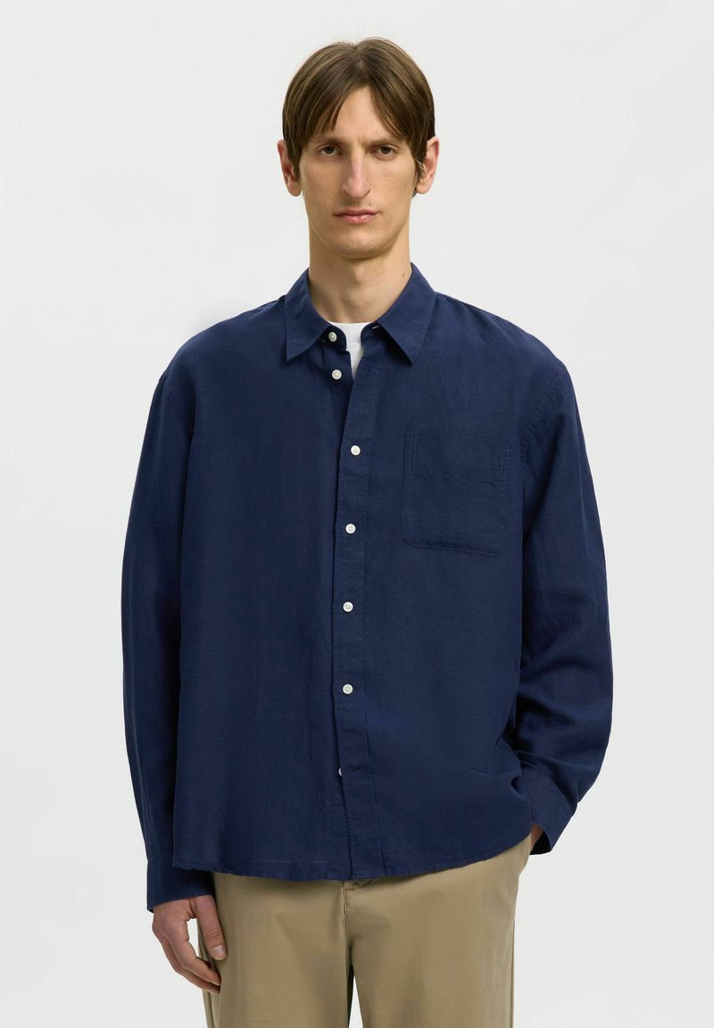 Homme aux cheveux bruns portant une chemise bleu marine boutonnée sur un sous-maillot blanc et un pantalon beige, debout devant un fond blanc uni.