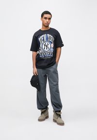 T-shirt gráfica preta dos New York Yankees com detalhes azuis, calças cinzentas afuniladas, botas bege e boné preto riscado. Fundo simples.