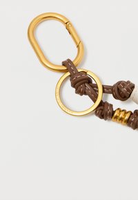 Kuldne karabiinide klipp, mis on kinnitatud võtmehoidjale, millel on pruuni nahknööri sõlmed ja kuldne metallpall valge tausta peal.