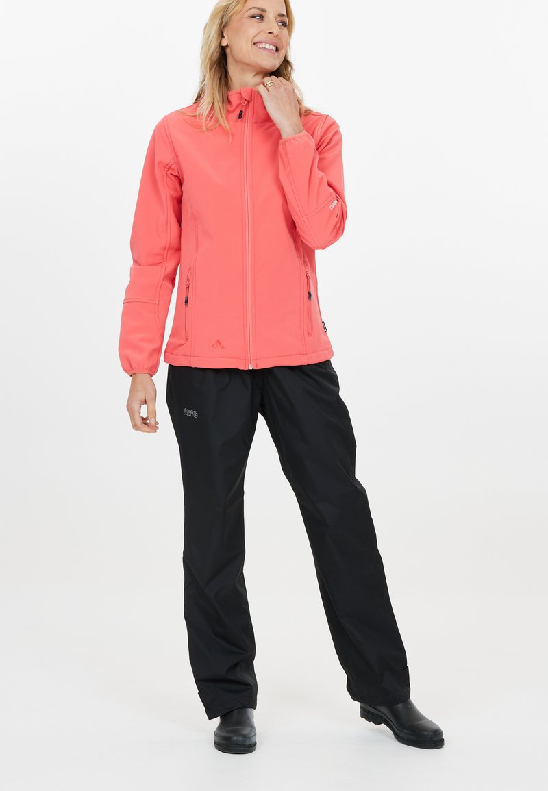Koraalkleurige softshell jack met een hoge kraag, ritszakken en elastische manchetten, gecombineerd met zwarte waterdichte broek en zwarte laarzen.