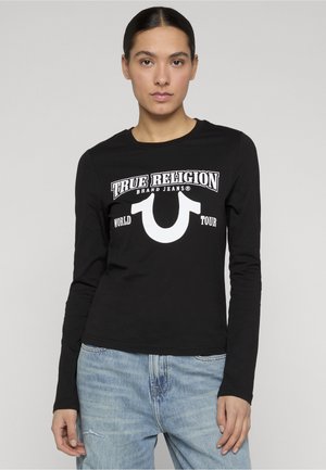 Femme aux cheveux foncés portant un t-shirt noir à manches longues avec le texte « True Religion Brand Jeans World Tour » et un jean bleu clair, debout devant un fond gris.