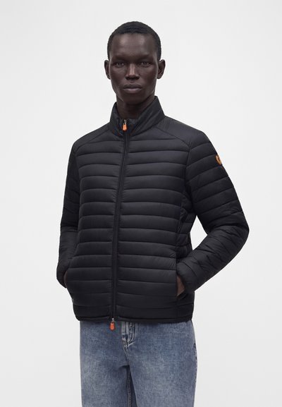 Homme portant une veste matelassée noire avec des curseurs de fermeture éclair orange et un jean bleu, debout les mains dans les poches devant un fond uni.