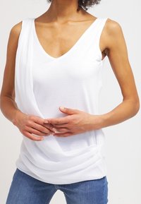 Top bianco senza maniche con scollo a V, caratterizzato da un tessuto drappeggiato sul frontale e una superficie liscia. Abbinato a jeans blu, con le mani posizionate sui fianchi.