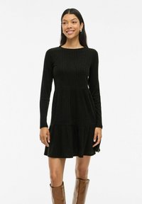 Robe noire côtelée à manches longues, col rond, jupe à volants et coupe décontractée. Présente un tissu texturé et un ourlet évasé.