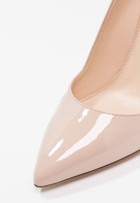 Chaussure à talon haut en cuir verni beige brillant avec bout pointu et coutures visibles le long du bord intérieur.