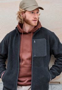 Schwarze Fleecejacke aus strukturiertem Stoff, mit einem Reißverschluss-Brusttasche und grauem Hoodie darunter; trägt eine beige Mütze.
