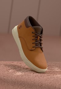Botín de cuero marrón hasta el tobillo con punta redondeada, detalles en marrón y suela color crema. Los cordones y el logo son visibles en el costado.